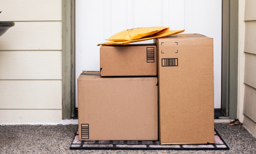 Three brown cardboard boxes and two yellow padded envelopes are stacked on a doormat in front of a white door, suggesting a home delivery.