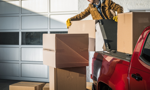 A person loads large cardboard boxes onto a red truck in front of a garage. They wear a brown jacket and yellow gloves, conveying a sense of activity.